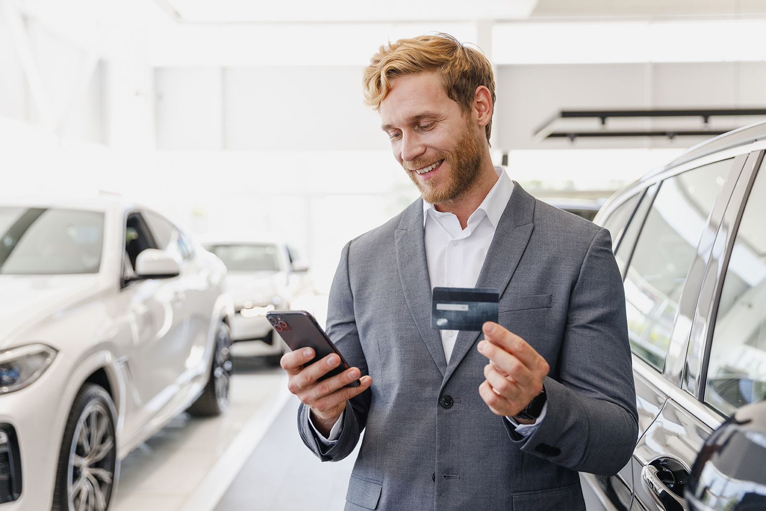 Car salesperson in a gray suit checks his phone while holding a credit card in a bright showroom with cars on display