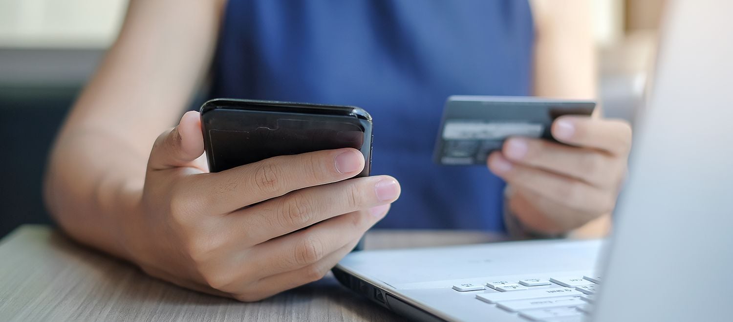 Person holding a smartphone in one hand and a credit card in the other, with a laptop on the desk for online shopping.