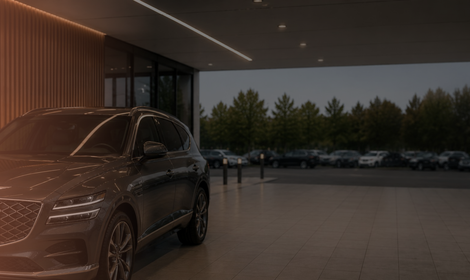 Front-left view of a dark SUV in a showroom with warm orange glow on the left and a row of cars outside under trees in the background, suggesting a dealership setting.